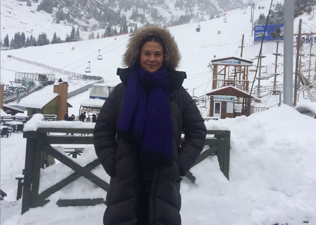 Photo shows a person dressed in a long grey parka and dark blue scarf, standing in front of a wintry scene at a ski hill.