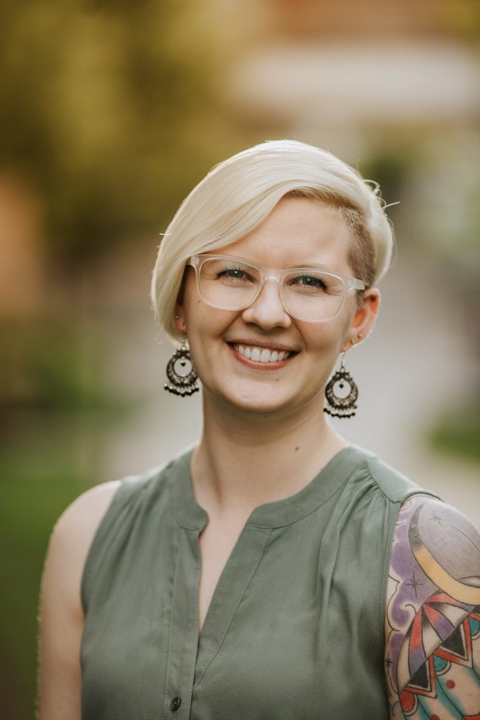 Image of a smiling woman with glasses and short blond hair, wearing big dangly earrings. She is wearing a moss green sleeveless shirt and has a colourful tattoo on her left shoulder